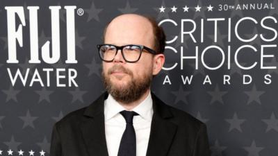 a man wearing a black tuxedo, he has a brown beard and black spectacles. He is stood in front of a black billboard advertising for Fiji Water and the Critics Choice Awards