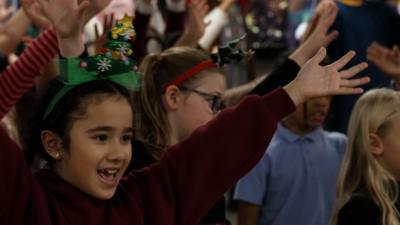 A girl wearing a Christmas Headband, waving her hands in the air whilst singing a song.