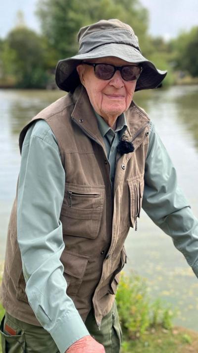 Peter Byers wearing a wide-brimmed hat, sunglasses and outdoor clothing stands beside a tranquil fishing lake holding a mobility aid.