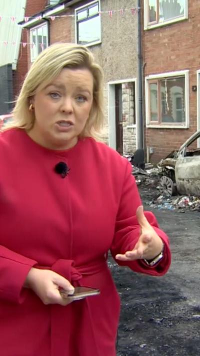BBC correspondent Sara Girvin weating pink coat and standing in front of street with debris and burnt car in the background