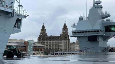 HMS Prince of Wales docked in Liverpool. The ship has a bridge where the time lapse footage of its arrival was filmed from. The deck is empty. Liverpool's Royal Liver building can be seen in the background