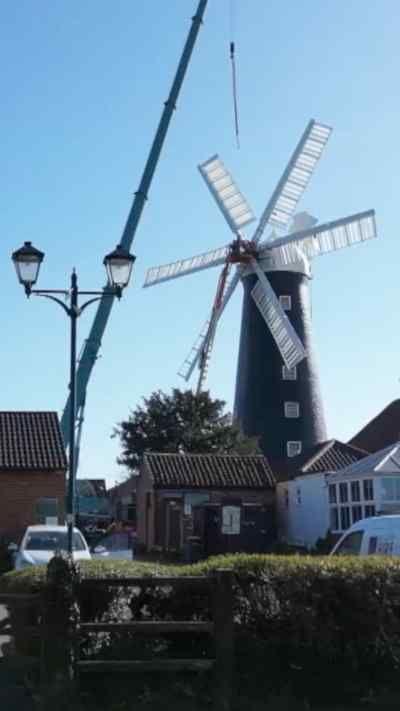 A large traditional windmill stands in the centre of the scene as a crane works beside it. Surrounding buildings, parked vehicles, and a clear blue sky frame the setting.