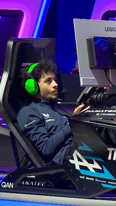 Young man with bright green headphones sitting in gaming chair in front of a screen driving a gaming wheel
