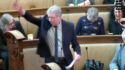 A screengrab taken from a live stream of a Hull City Council meeting. Alan Gardiner, wearing a dark blue suit, white shirt and blue tie is standing with his arm in the air. He has grey hair and glasses.