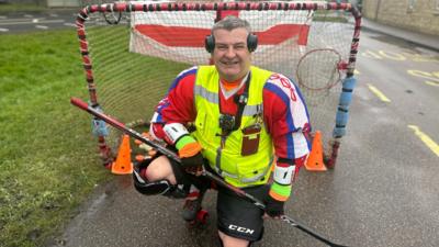 Paul Hinson, wearing roller hockey equipment, knees down in front of a goal