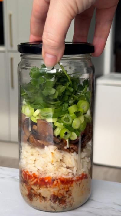 A jar filled with rice, mushrooms, spring onion and sauce on a marble counter top