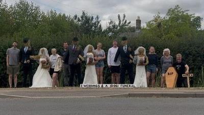 A line-up of eight real people surrounding three scarecrow couples who are wearing wedding dresses and suits complete with black hats, at the far right is a small wooden coffin bearing the letters 'RIP'. A sign next to that says 'Alfie' who was the family dog that died last month. A longer white sign on the ground reads 'Three weddings and a (dog) funeral'.