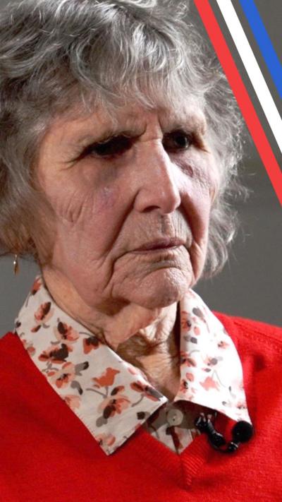 Olga Henderson is sat in a room with a plain grey background. Light is hitting her face and curly grey hair from the window as she looks emotional while speaking to an off-camera reporter during an interview about being a child prisoner in a Japanese camp. She is wearing a red jumper with a floral shirt beneath and dangling earrings.