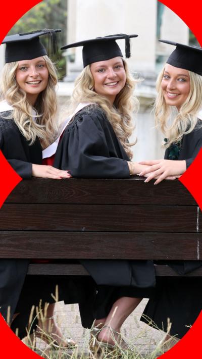 A set of triplets sat on a bench, smiling, and wearing a cap and gown.