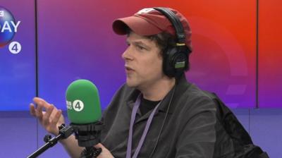 Jesse Eisenberg wearing red cap and headphones speaking into a mic in radio studio