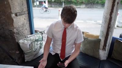 Student wearing a white shirt and red tie looks down at his mobile phone screen