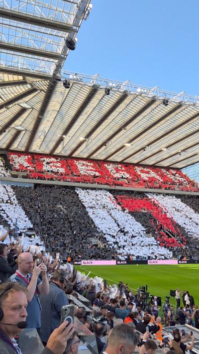Crowds at St James' Park hold red, white and black cards to create a large mosaic display across the stands spelling out 'Keegan' with a red number '7' below. The green pitch is visible further below and a clear blue sky above the stadium roof.   