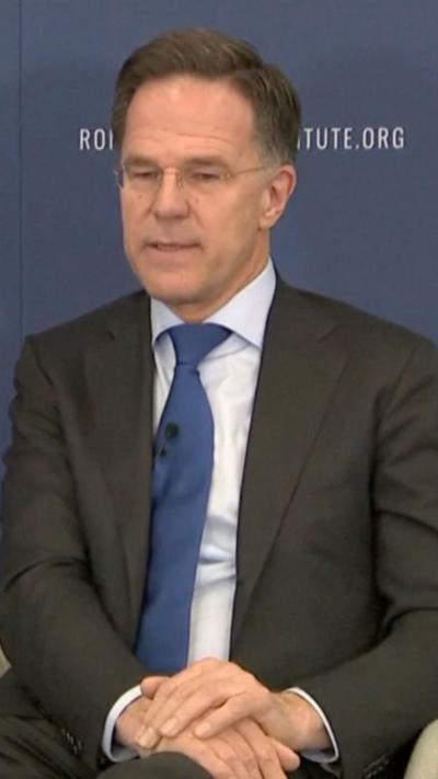 Mark Rutte wears a dark suit, purple tie and white shirt while seated with his hands folded, he has a pensive look to him.