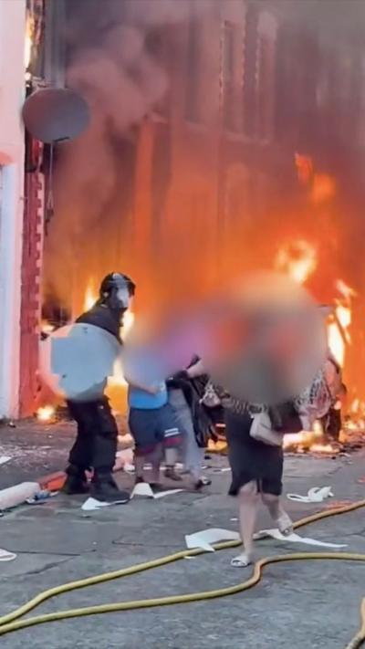 Police wearing face shields escort people on a street, as fire and smoke is seen behind them