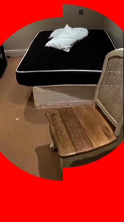 Image is a bedroom. in the background is a black mattress with a white pillow on top. In the foreground is a wooden chair.