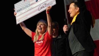 A female is holding onto a check with Elon Musk next to her.