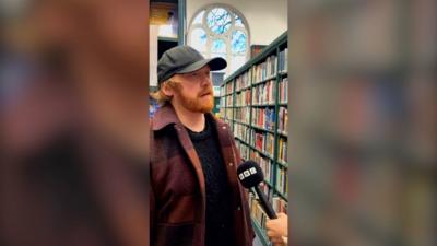 Rupert Grint stands in a library wearing a hat