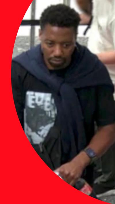 A man with curly black hair and a goatee moustache and beard. He wears a black T-shirt with a white picture printed on it, and a navy blue sweatshirt knotted over his shoulders.