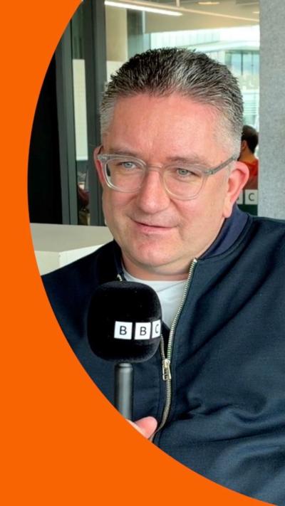 A man, Dan Finley, with grey hair and clear glasses, holding a mic.