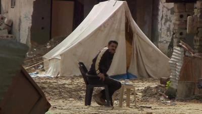 Man sits amongst rubble by tent