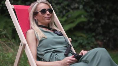 Lara Lewington sits in a deckchair. She is wearing sunglasses, holding a smart skipping rope and wearing a Sensate device.