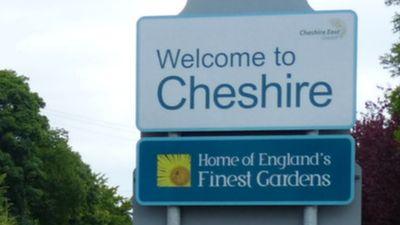 A white road sign with blue writing, which reads "Welcome to Cheshire", above a blue road sign with a yellow flower in a square and white writing which reads "Home of England's Finest Gardens"