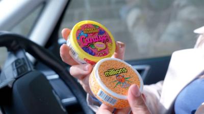 A photo of two small tubs of nicotine pouches being held in a reporter's hands. One is orange and has a picture of a smiling orange on the cover with loads of small oranges under neath the word Millions. It does not mention the word nicotine. The Candys pack is pink with colourful gummy bears on it. It says "Sweet nicopods".