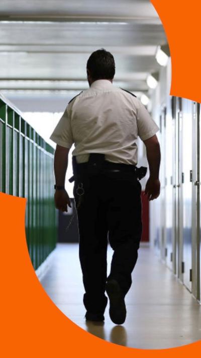 A prison guard walking through a corridor. Doors on the right, green fencing on the left.