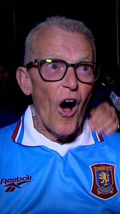 Villa fan shouting into camera. Mouth open, wearing a retro Aston Villa shirt and dark rimmed glasses