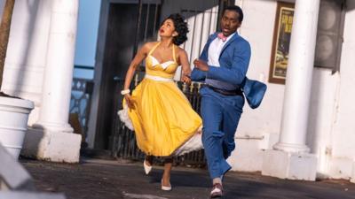 A man in a blue suit and a woman in a yellow dress run from a doorway