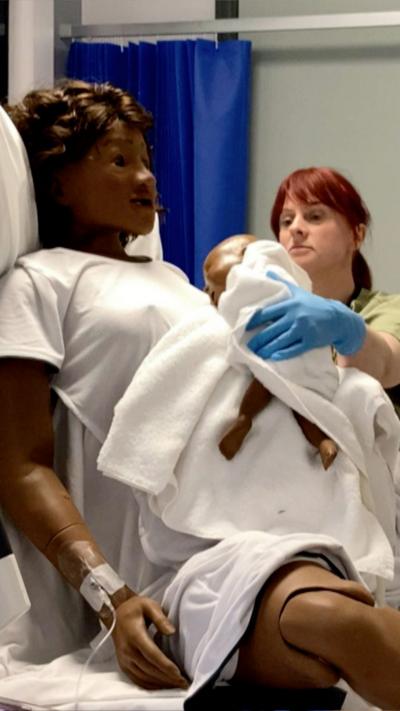 A mannequin resembling a woman in labour is attended to by a woman who holds a baby to its chest