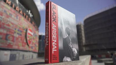 A copy of the book Black Arsenal standing up on a plinth with the Emirates Stadium in the background