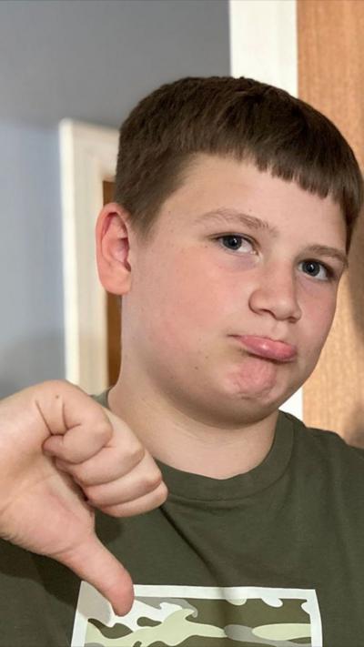 A boy with sort brown hair stands making a thumbs down sign