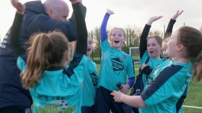 a group of girls wearing football kit cheering together with their coach while standing on a grass football pitch