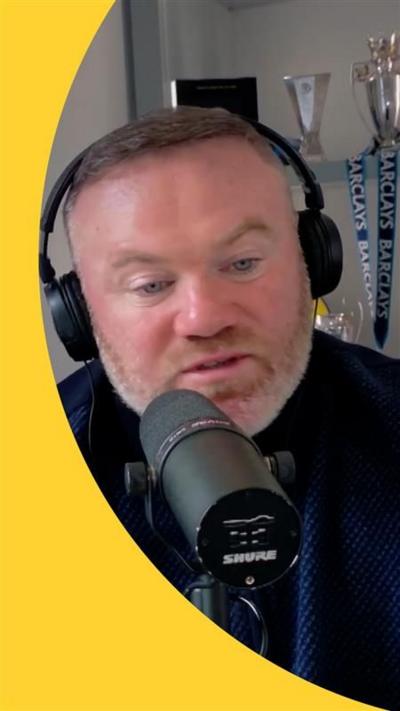 Wayne Rooney speaks into a mic while wearing headphones. Trophies are on display behind him 