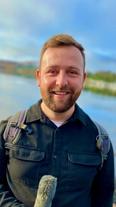 Jamie McDonald holds a microphone, behind him is a loch on the Isle of Skye