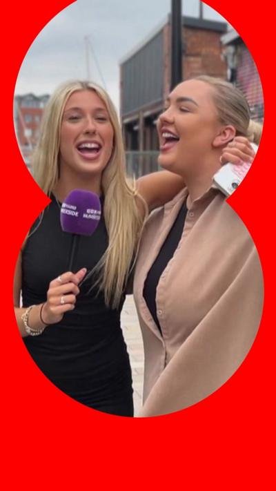 Two women, with their arms around each other, sing in the street. One holds a purple mic