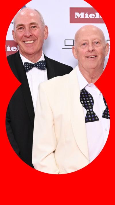 Race Across The World finalists and brothers Melvyn and Brian Mole on the red carpet at the National Television Awards. Brian on the left is wearing a black suit jacket and white shirt and done up polka dot bow tie. Melvyn on the right is wearing a white suit jacket with an undone polka dot bow tie