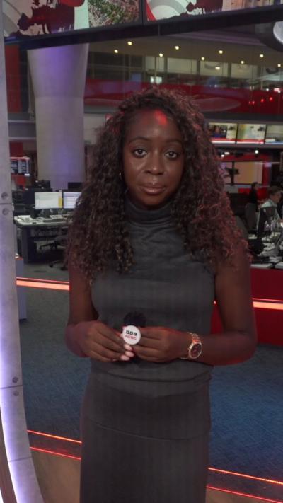 A woman with long, brown curly hair wearing a brown dress talks to the camera