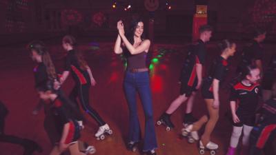 Presenter Vicky Hawkesworth claps in the middle of a roller rink, surrounded by kids setting off skating