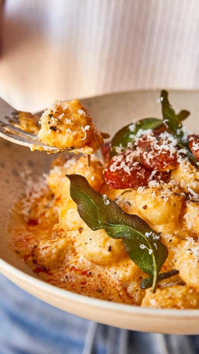A person holding a white bowl with butternut squash gnocchi in one hand and a fork in the other