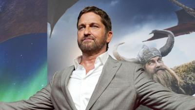 Gerard Butler wearing a grey suit jacket and a white shirt. He has dark hair and facial hair. Behind him is a poster for the film.