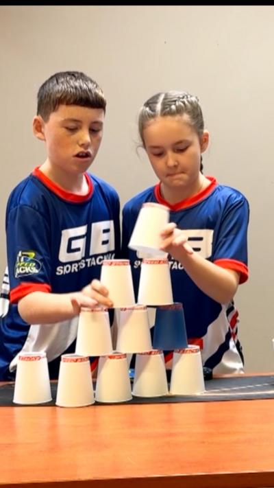 Two children - a boy with short brown hair and a girl with blonde hair pulled back behind her head - are stacking white cups in a pyramid. They are wearing dark blue sports tops with "GB sport stacking" written on them in white.