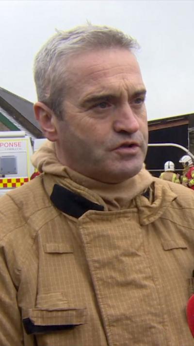A firefighter with short grey hair, looking at the camera. There are a number of fire engines behind him.