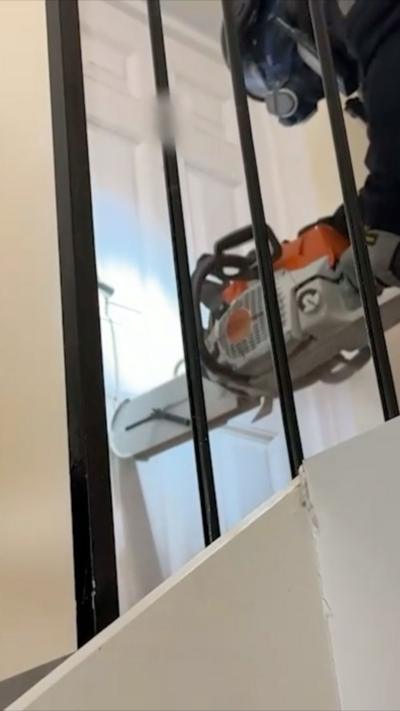 A man in a mask using a chainsaw on a white door behind some black railings