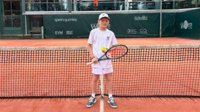 A young boy in tennis whites holding a racket and ball on a tennis court.