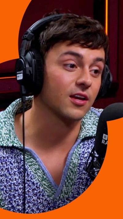 Tom Daley speaks during an interview in a Radio 2 studio. He has short brown hair, and wears headphones and a green and blue knitted top