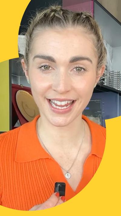 A woman with blonde hair pulled away from her face smiles as she talks to the camera. She wears an orange jumper