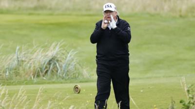 Donald Trump on Turnberry golf course wearing dark trousers, dark zip up top and white cap with 'USA' on the front. He is holding a golf ball and has his hands at either side of his mouth as he shouts back at reporters asking him questions.