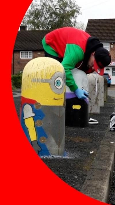 A bollard painted as a Minion, side view. A man behind it in red and green jacket is painting another one.
